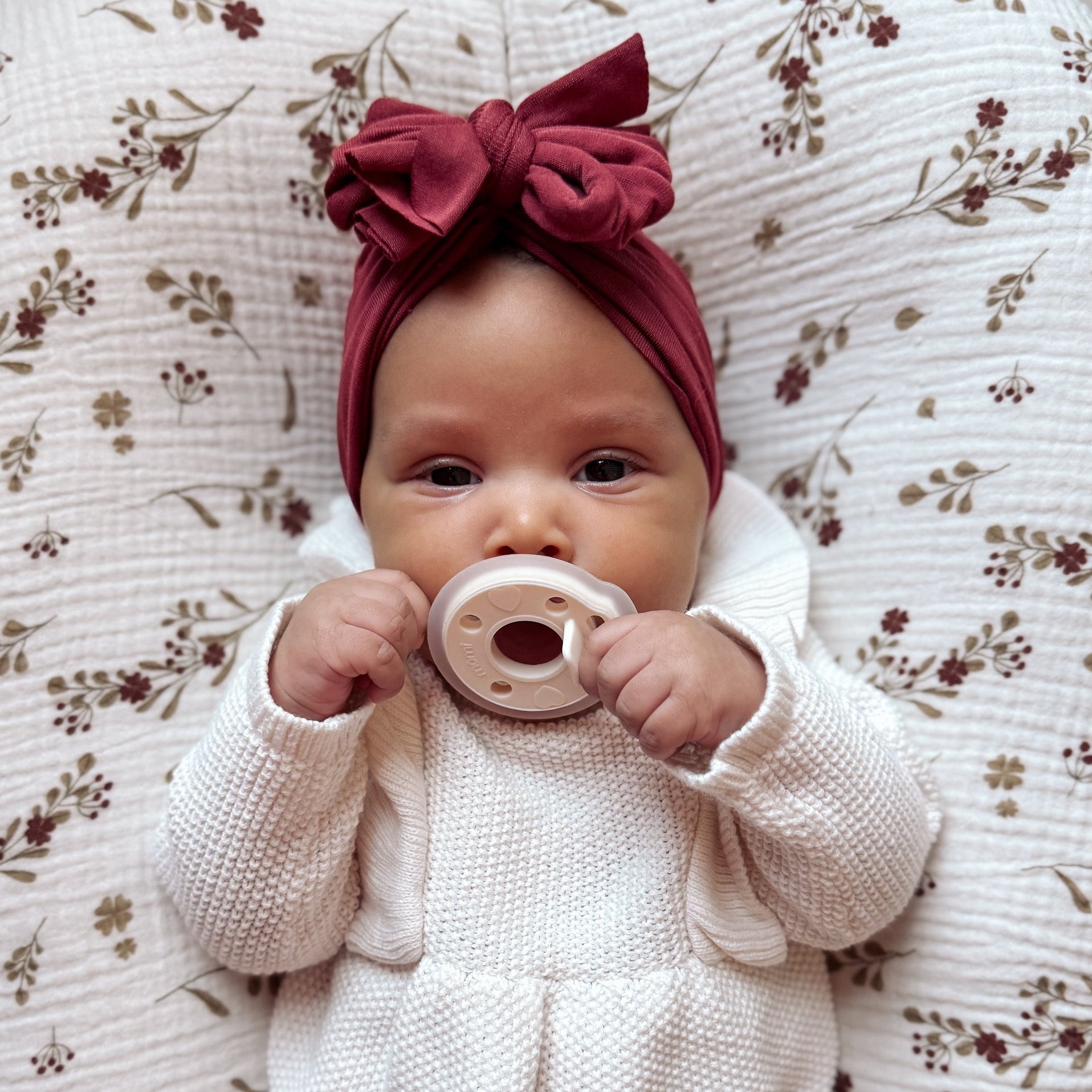 Baby in a white outfit with a red bow headband holding an antique-white "pearl" mōmi  pacifier on a floral-patterned cushion