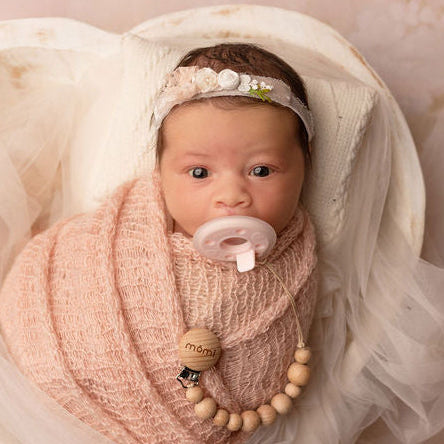 Newborn baby wrapped in pink blanket with mōmi soft pink pacifier and natural wood pacifier clip pacifier, lying in heart-shaped bowl on floral pink background